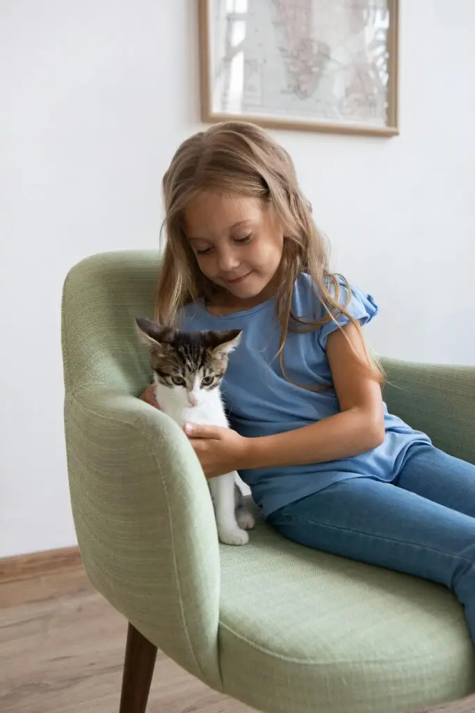 A young girl lovingly holds a kitten while sitting on a green chair indoors.